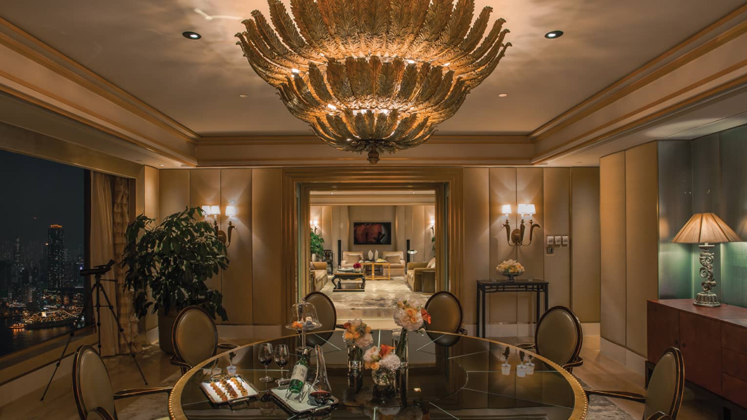 A luxurious dining room with a glass table and elegant chairs adorned with floral centerpieces. A large, ornate chandelier hangs from the ceiling.