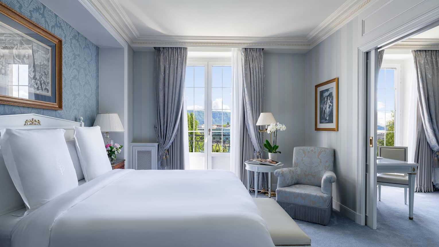 Luxury hotel room with soft blue undertones, featuring a king bed, bench, upholstered armchair, small table and double doors opening to a balcony with city view