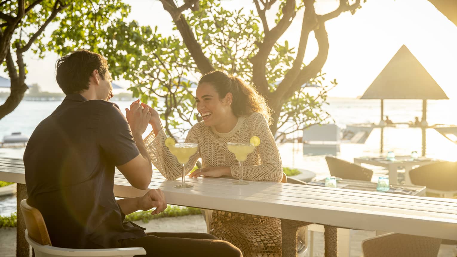 Laughing couple enjoys cocktails at outdoor beachside restaurant surrounded by greenery