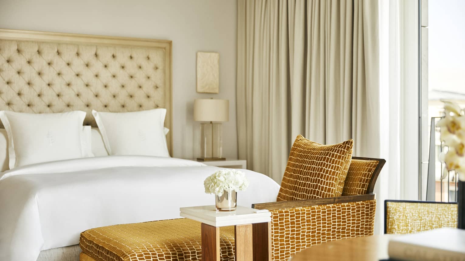 Close-up of a king bed, tufted headboard, nightstand with lamp, tan chaise lounge with matching pillow, small side table with white flowers and beige curtains