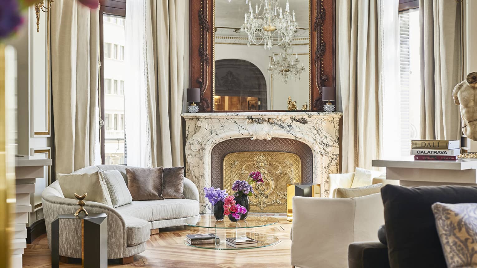 Luxury seating area with a curved sofa, ornate marble fireplace with gold accents, large mirror, crystal chandelier and floral arrangement on a glass table