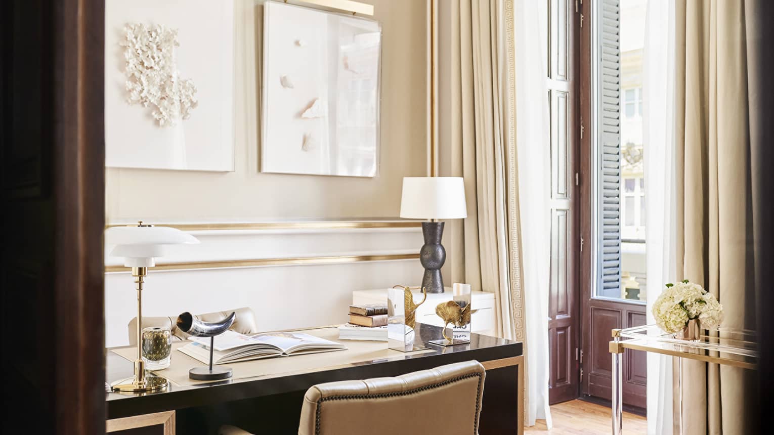 Luxury hotel suite office with beige walls, gold accents, desk with decorative objects, books and a modern lamp, next to tall windows with cream curtains