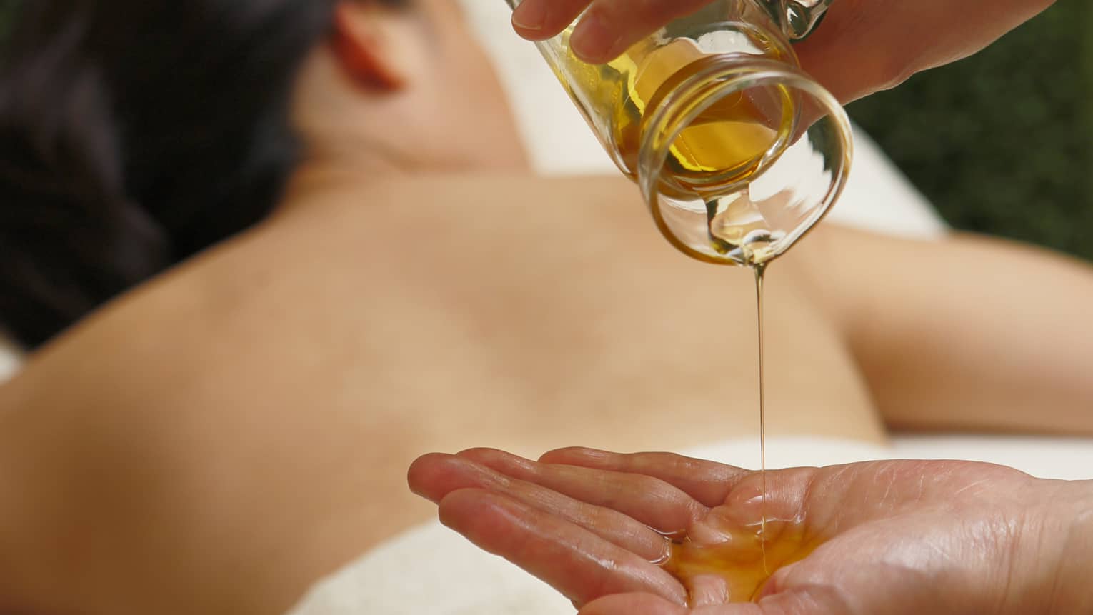 Spa staff pouring massage oil from glass jar in hand over woman lying on table