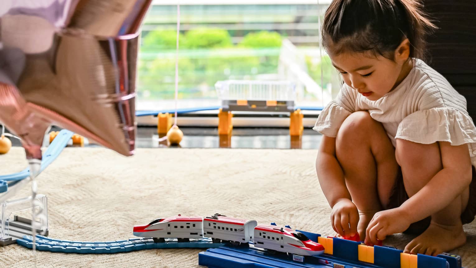 Young girl plays with train, large window and star balloons in the background
