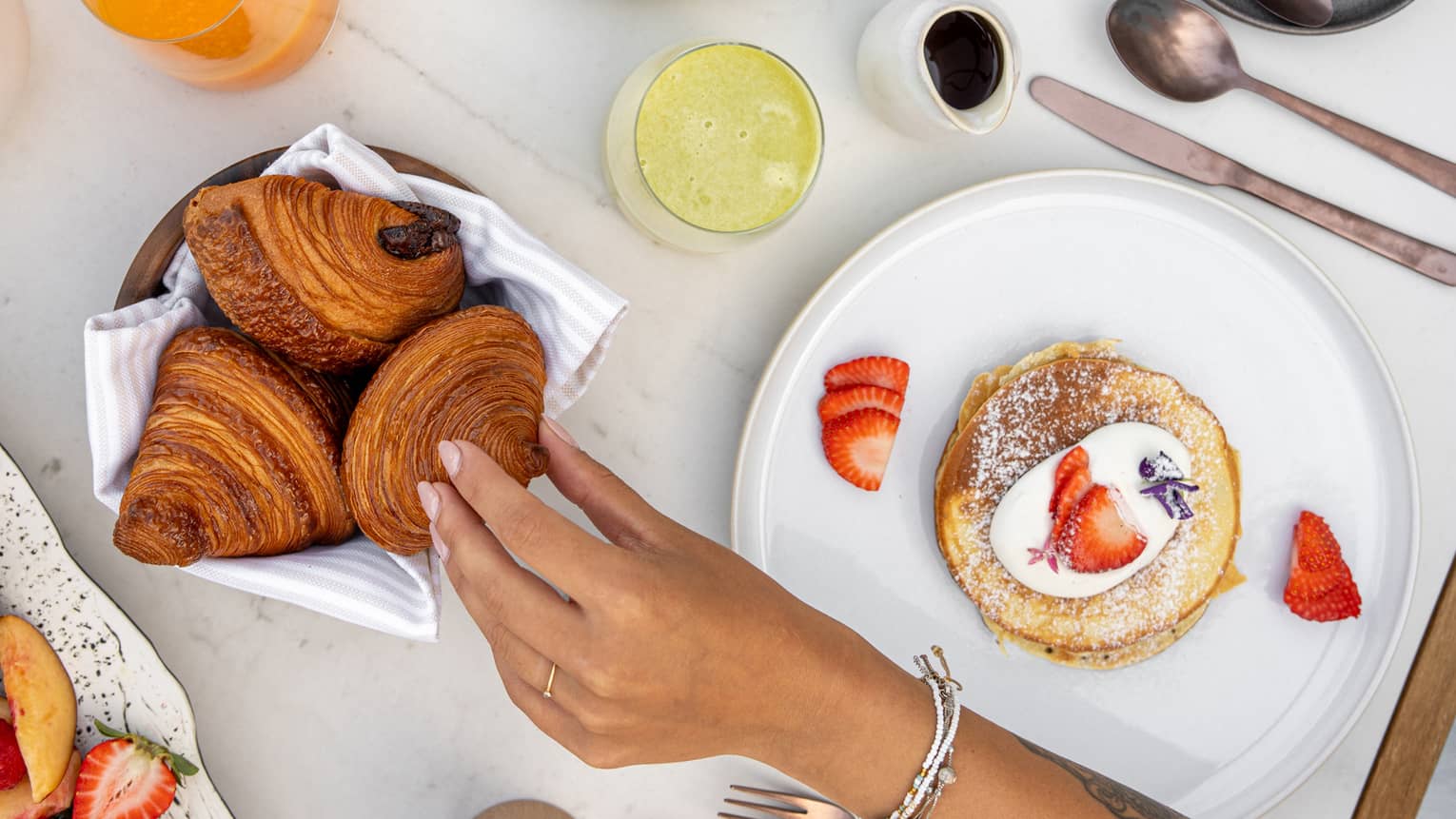 An array of plated breakfast food items, as well as coffee and juice, set out on a table. A person reaches for a pastry item, while another person uses utensils to cut into the food on their plate.