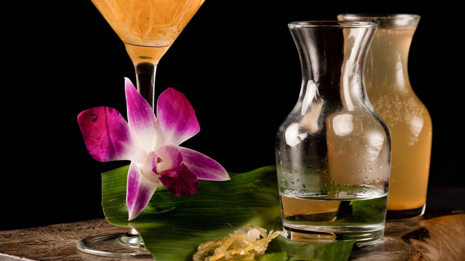 An orange martini glass on a wooden table surrounded by several pitchers of alcohol.