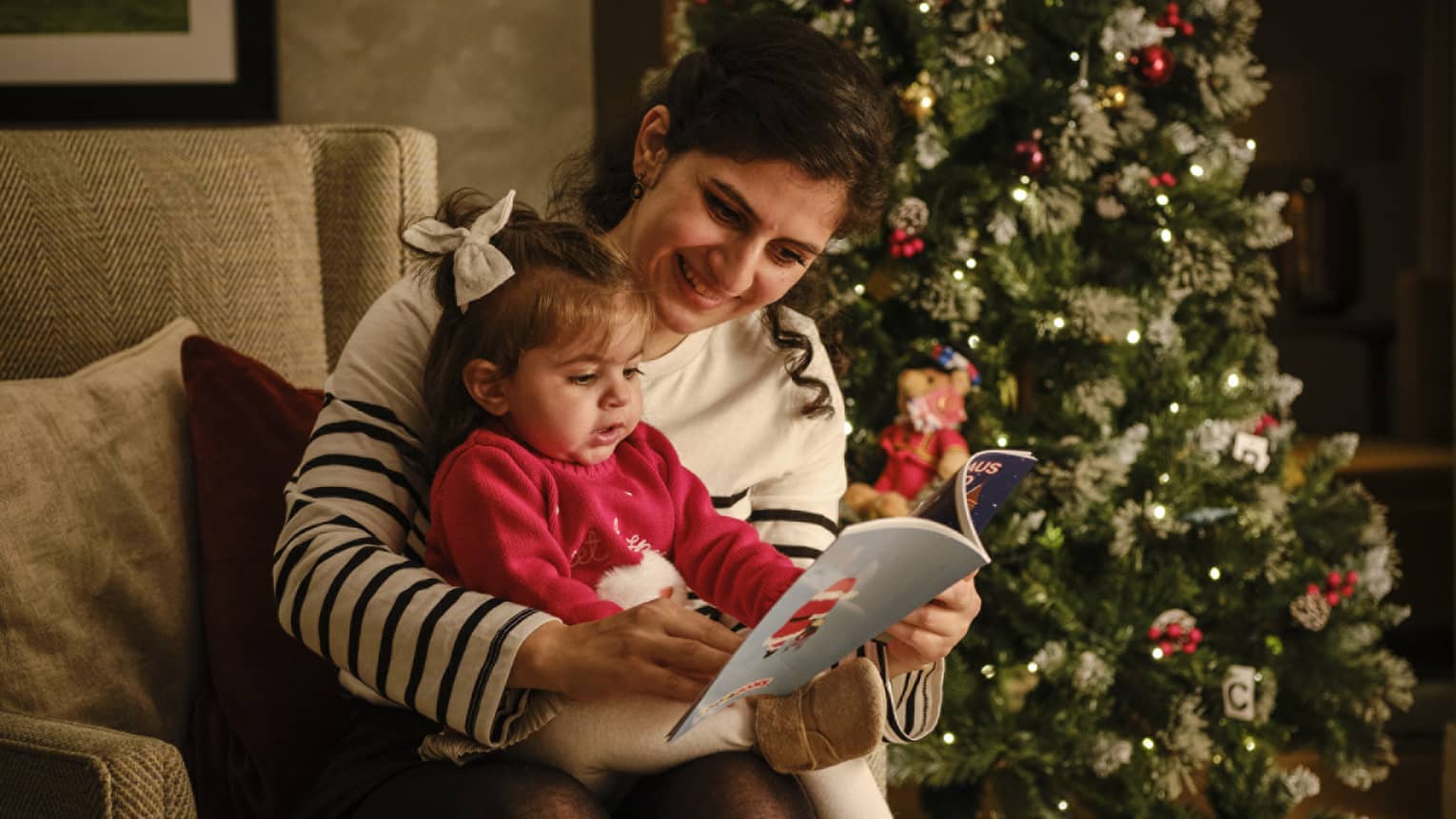 A woman reads a book to a small child while sitting in front of a decorated Christmas tree