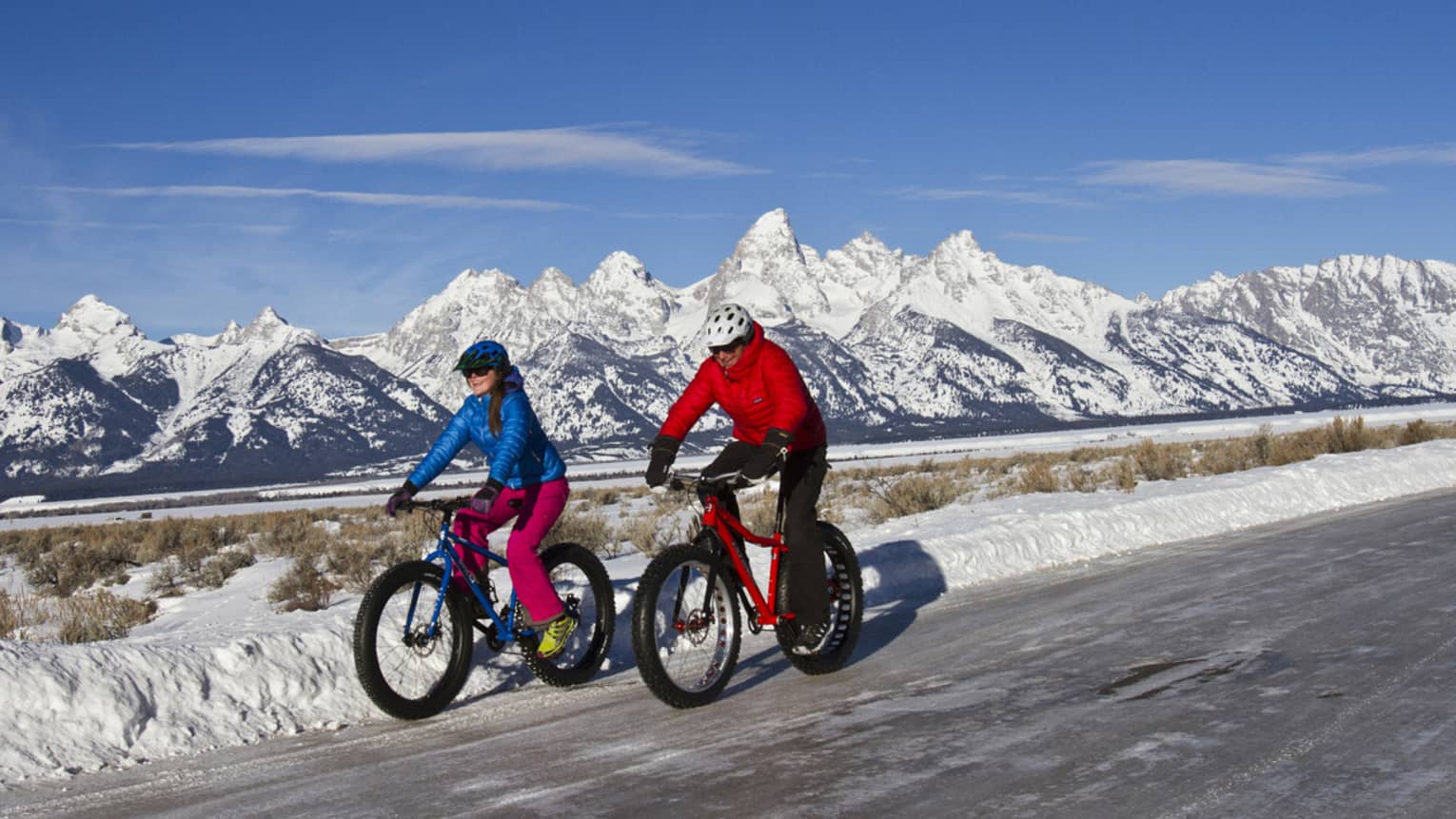A man and woman bike down a path, snow-capped mountains and blue sky in the background