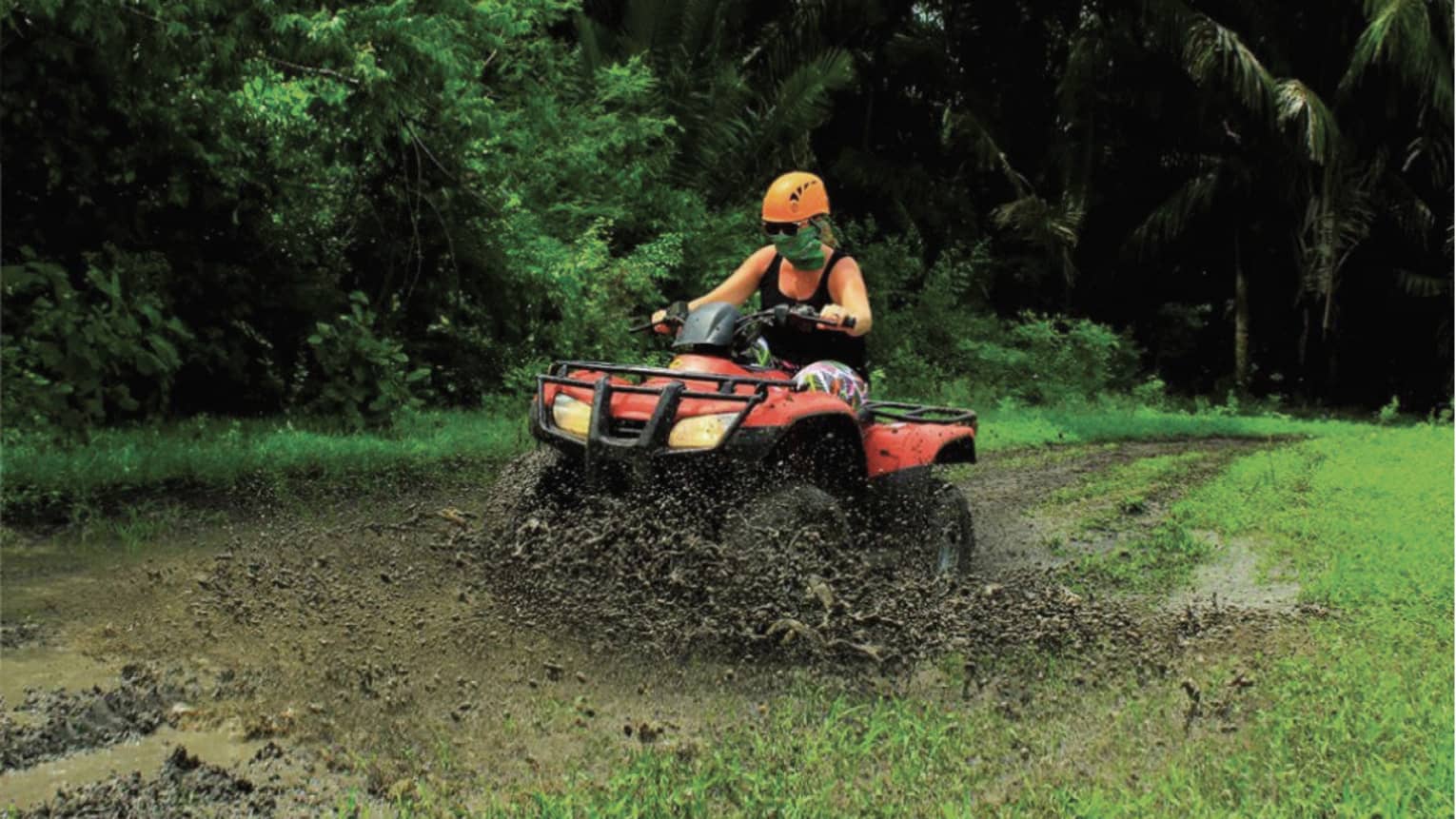Person wearing a black tank top and bright orange helmet rides through the mud on a red ATV