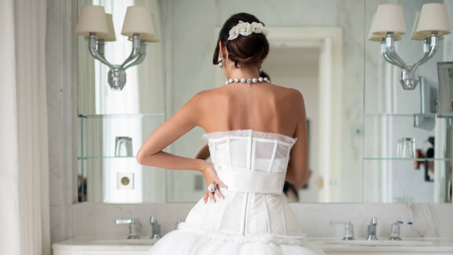 Back view of bride standing at vanity mirror in couture strapless wedding gown, hair up, hand on hip