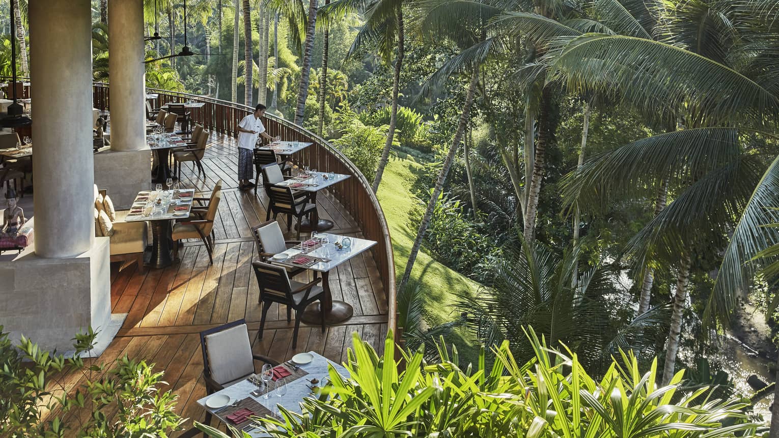 Looking down at curved wood patio of Ayung Terrace outdoor dining room, tables and chairs overlooking palm trees, forest