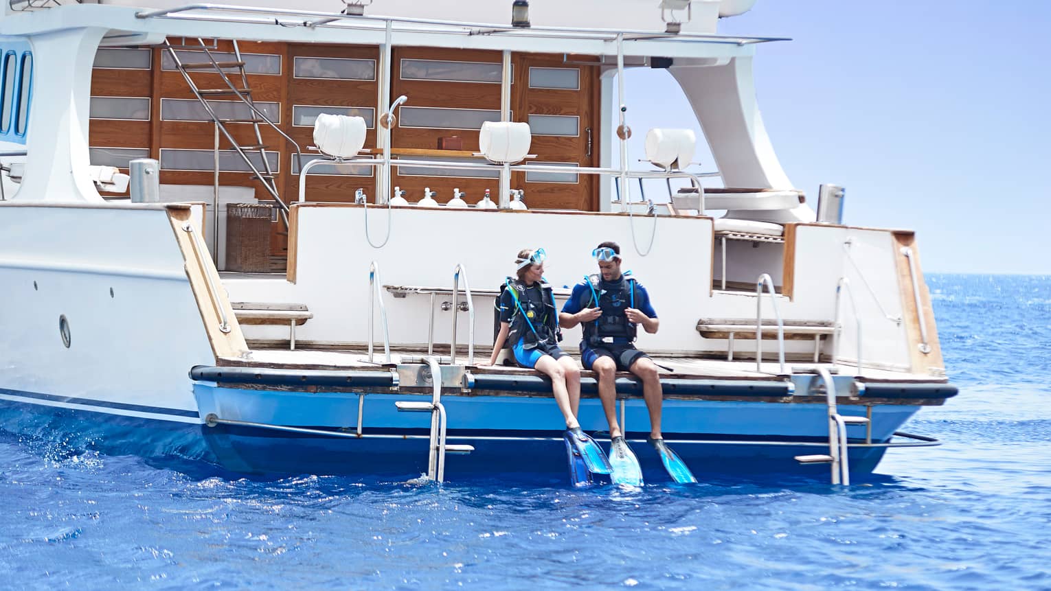 Man and woman wearing scuba diving gear sit on back of white yacht