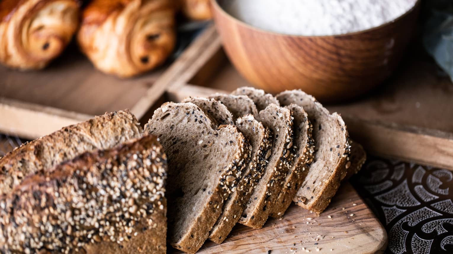 Loaf of rye bread sliced half sliced half whole with a bowl of flour and a couple croissants in the background