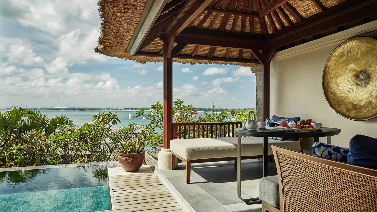 Close-up of Deluxe Villa patio corner with swimming pool, plant, white bench under thatched roof, ocean view