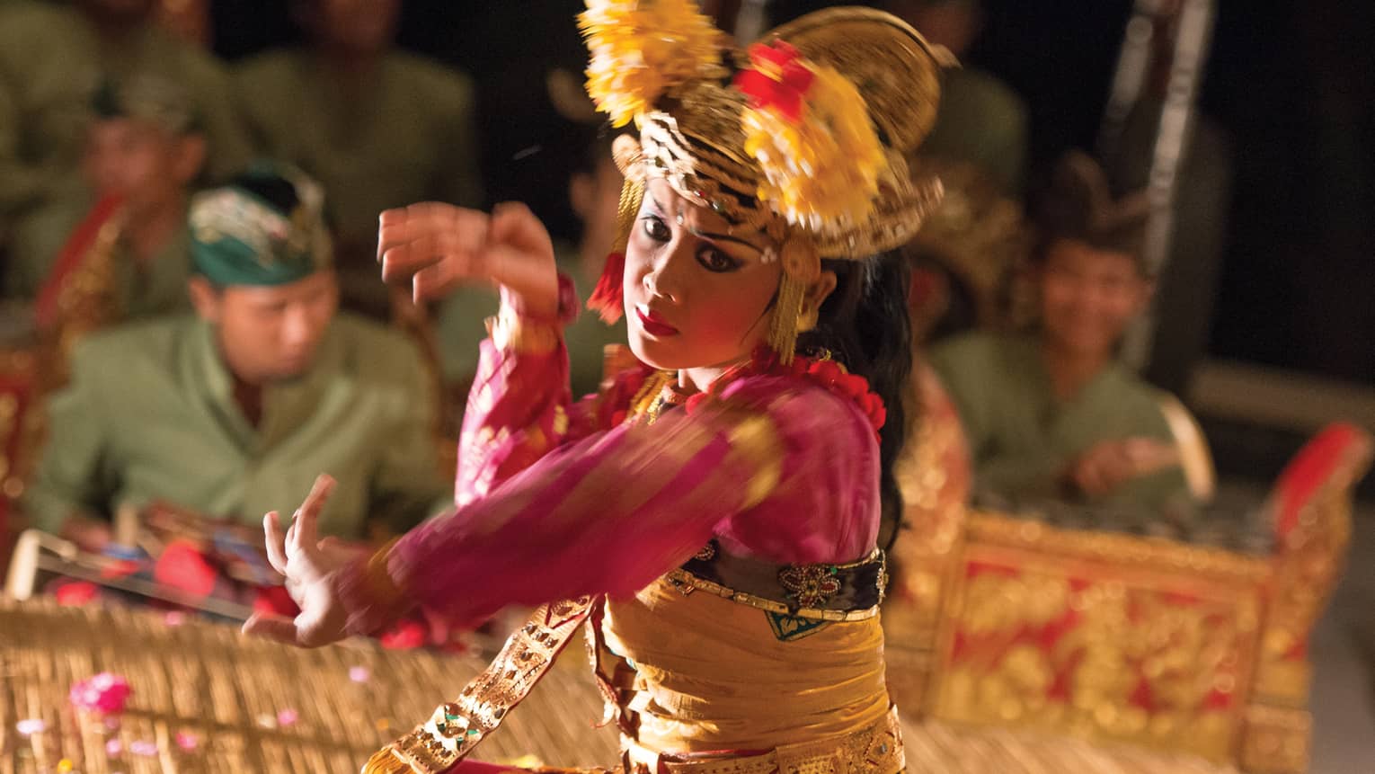 Close-up of a Balinese dancer in a golden headdress and bright clothing; in the background musicians play the gamelan. 