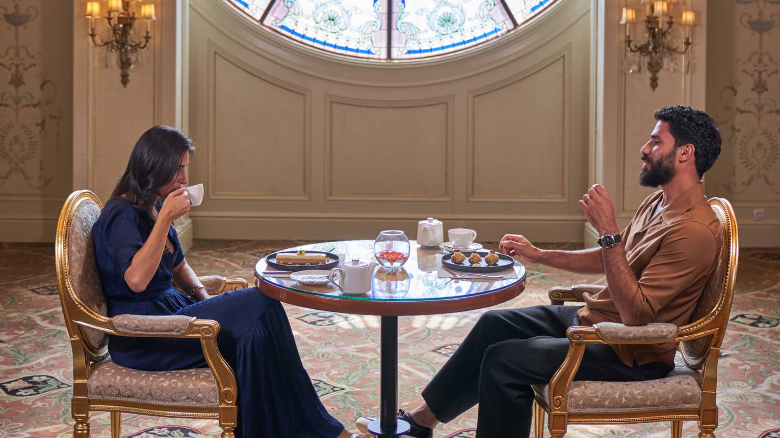 Couple sits across from each other sipping tea at small round table in front of large round window adorned with blues and greens