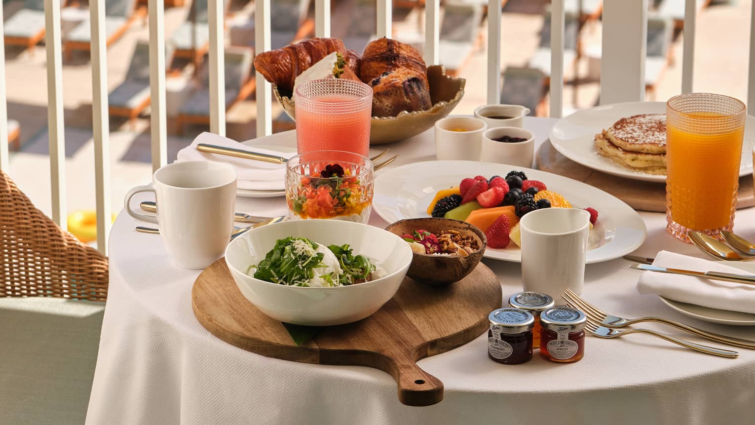 A small round table covered with a white table cloth holds a variety of breakfast items and beverages, all on an outdoor balcony overlooking the beach