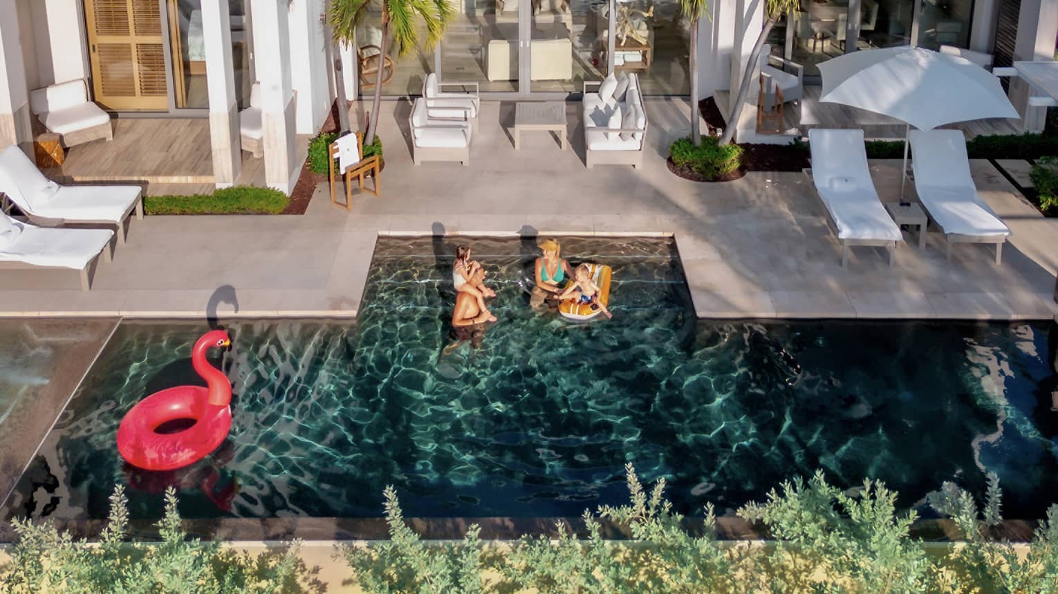 A young family wades with inflatables in a pool surrounded by white lounge furniture, umbrellas and palm trees.