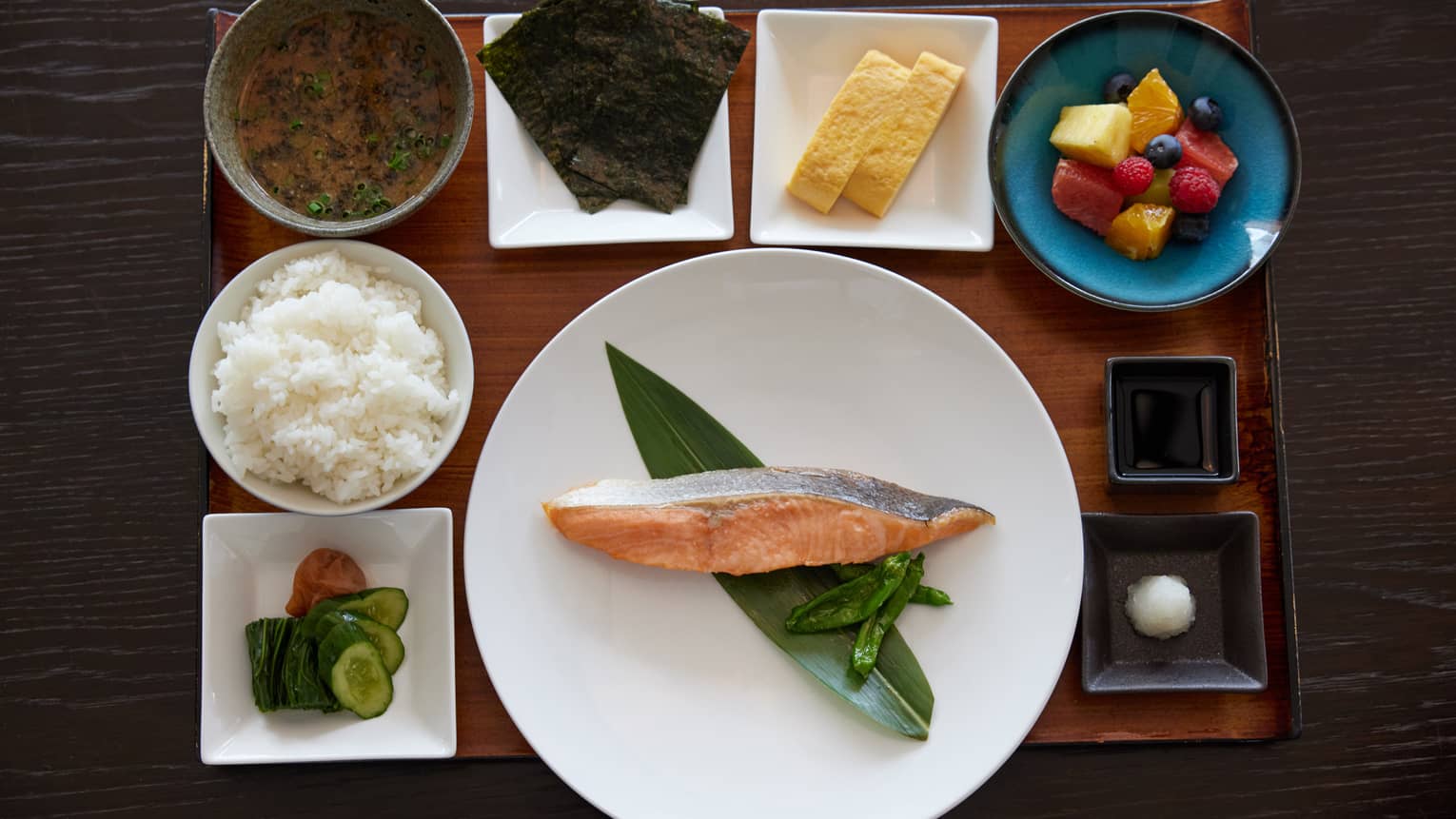 On wooden tray in dishes of various shapes and sizes, an alluring spread of food items, including fish, rice and fresh fruit.