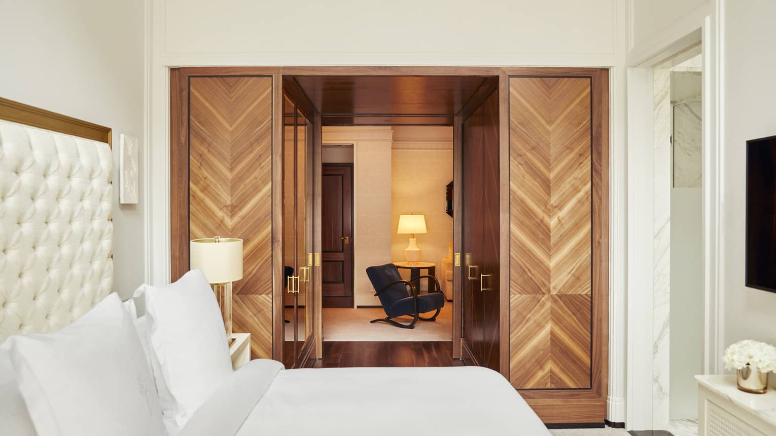 Luxury hotel suite bedroom with a king bed with tufted headboard and double wooden doors leading to a cosy lounge area with a dark armchair and table lamp