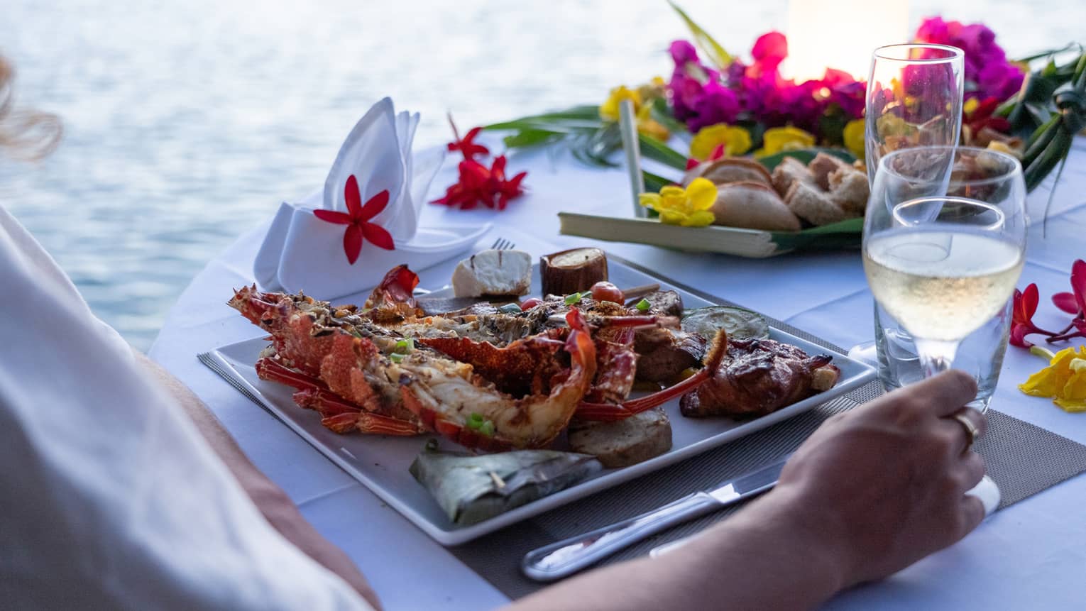 Close-up of a table laid with tropical flowers, wine and food, including shellfish, breadfruit and wrapped banana leaves.