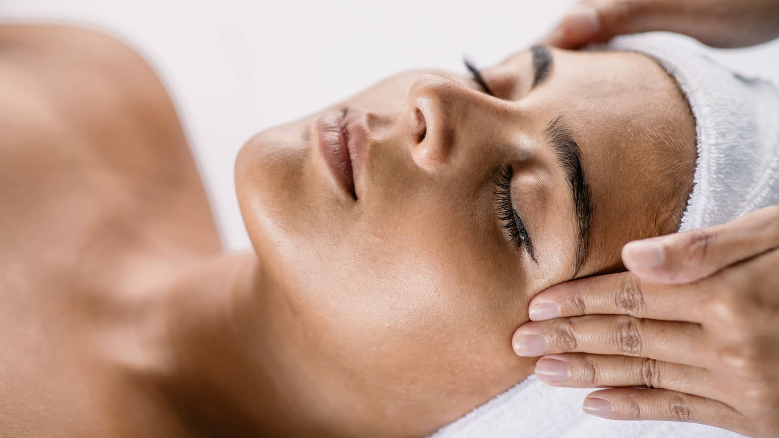 Hands massage woman's temples as she closes her eyes, spa towel around hair