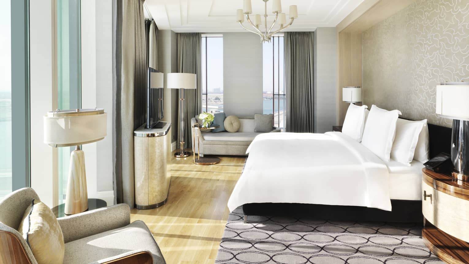 Executive Suite bed with white linens under chandelier, sunny corner windows, grey loveseat sofa and chair