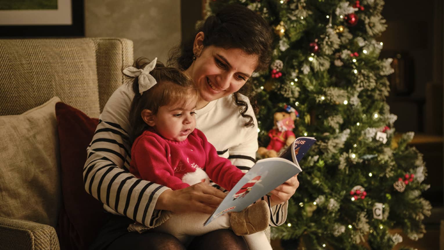 A woman reads a book to a small child while sitting in front of a decorated Christmas tree
