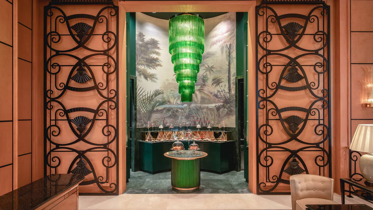 View through an ornate orange doorway of a tiered green chandelier hanging above tables of desserts covered by glass domes.