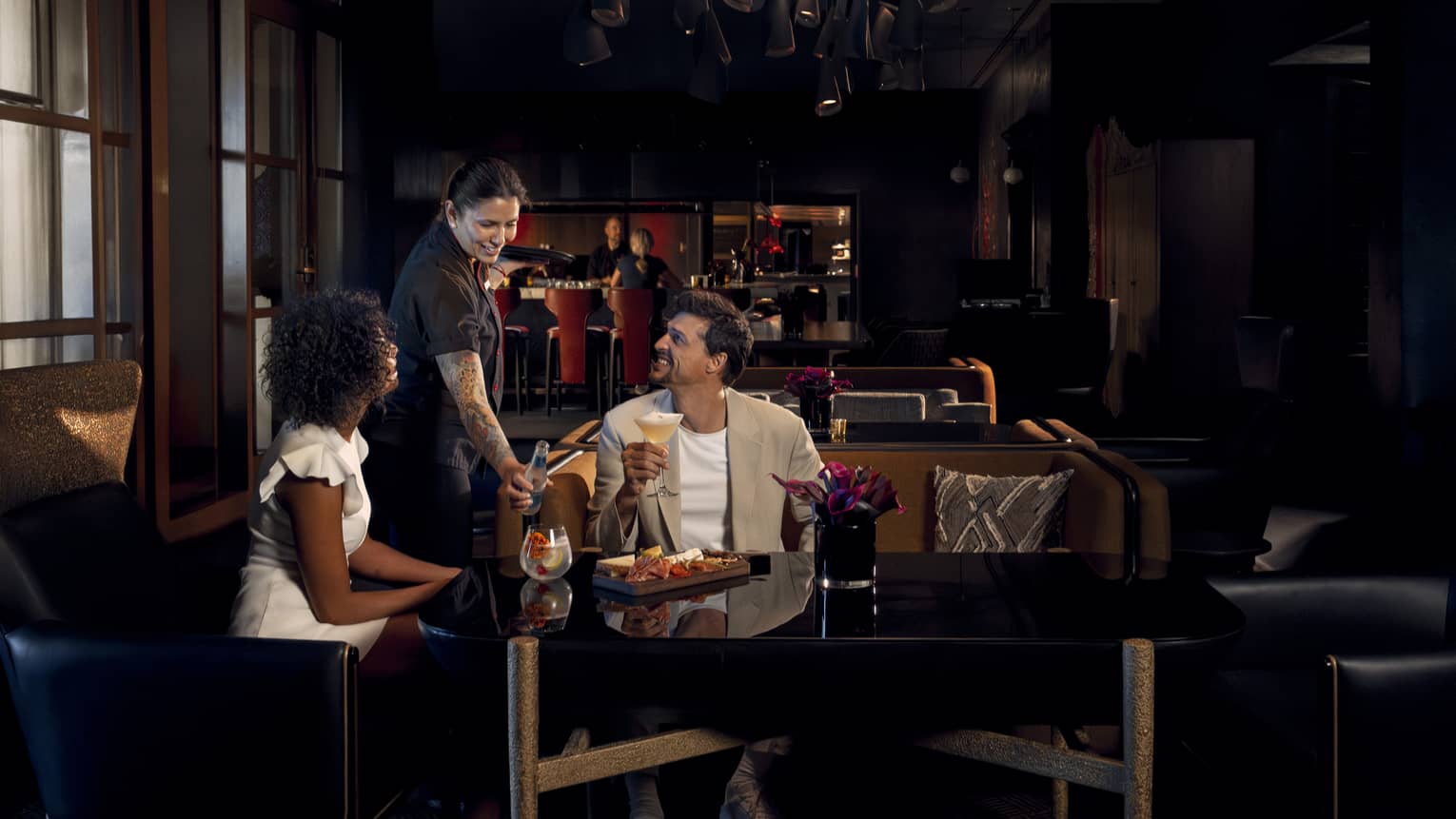 Two people wearing light-coloured cocktail attire sit on benches around a black table as a waitress brings them beverages