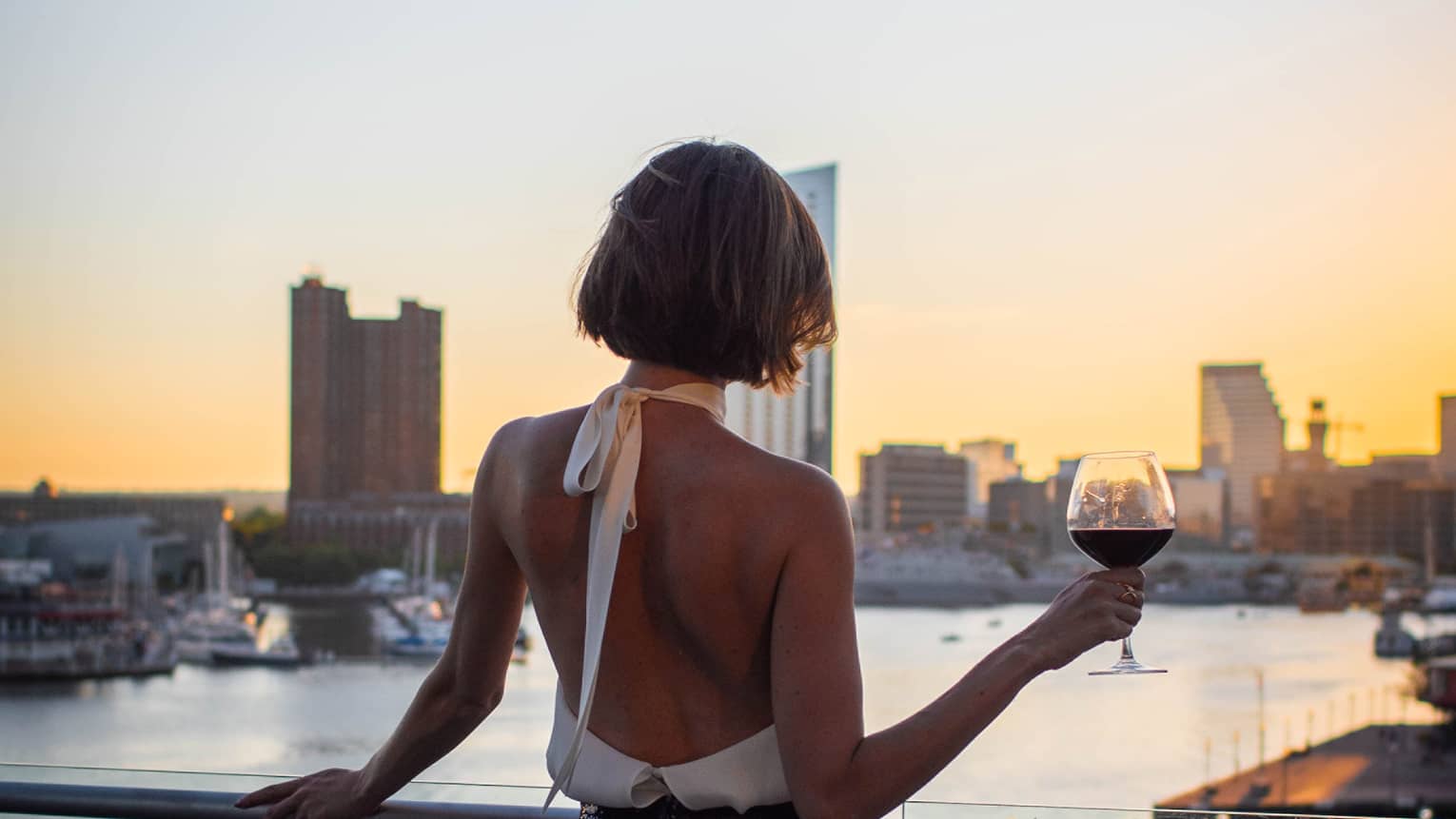 Person with bob haircut wearing a white halter-neck top and dark skirt stands silhouetted at sunset as they hold a glass of wine and overlook a city harbour