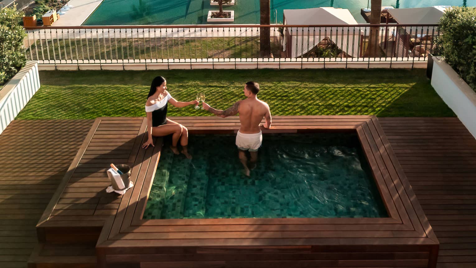 Two people in a small square pool in an outdoor setting. A larger pool surrounded by lounge chairs is in the background, as are ocean and mountain views beyond that. Verdant lawns and trees surround the pool areas.