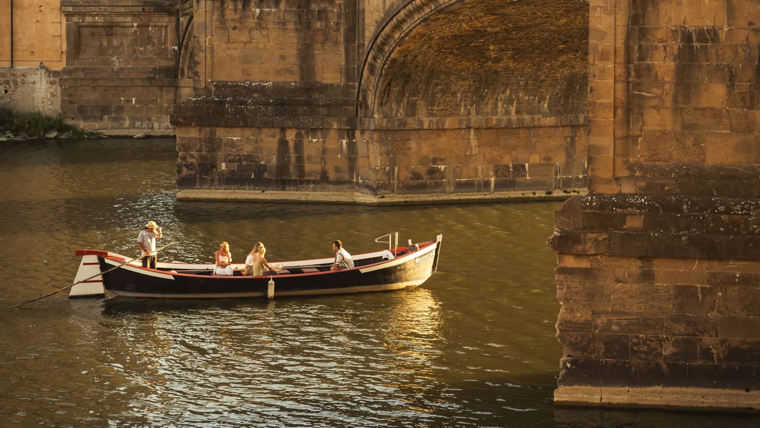 A barchetto, an ancient boat, with a few people on it are leisurely making their way under a crowded stone bridge.