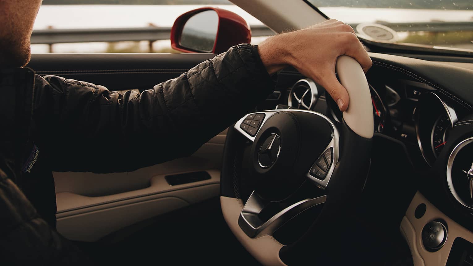 Passenger's view of driver gripping the wheel of a Mercedes convertible, looking away at a lake and distant mountains.