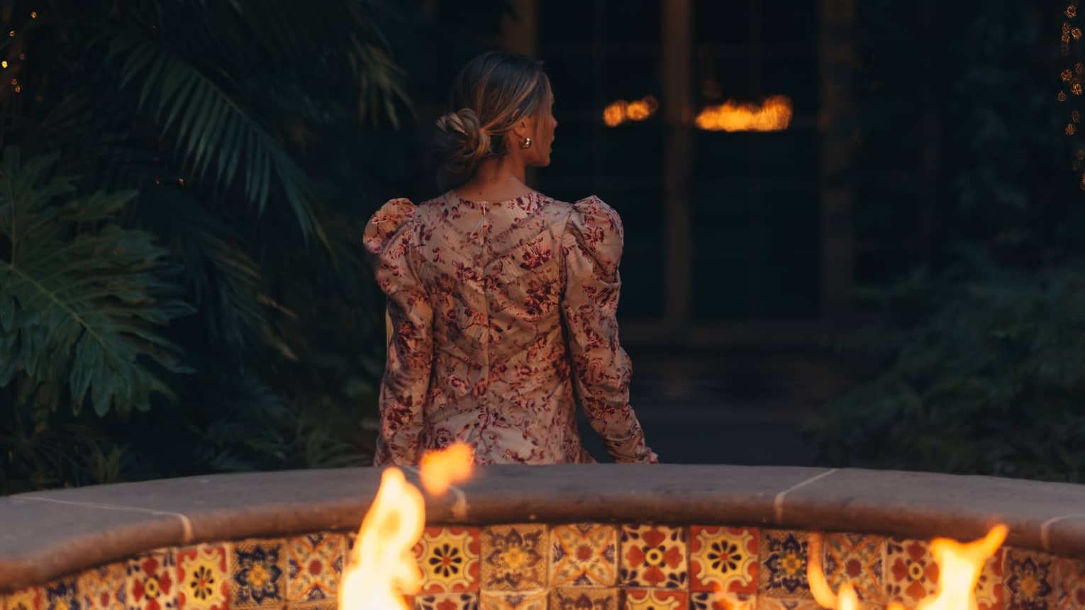 Rear view of a person in an outdoor evening setting standing beside a tiled firepit and facing a foliage-covered wall and arched doorway