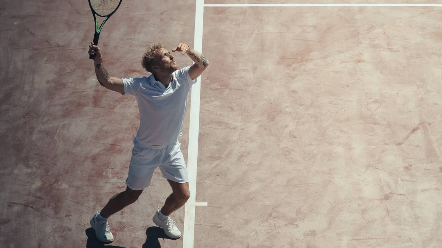 Man in white outfit playing tennis on reddish clay court