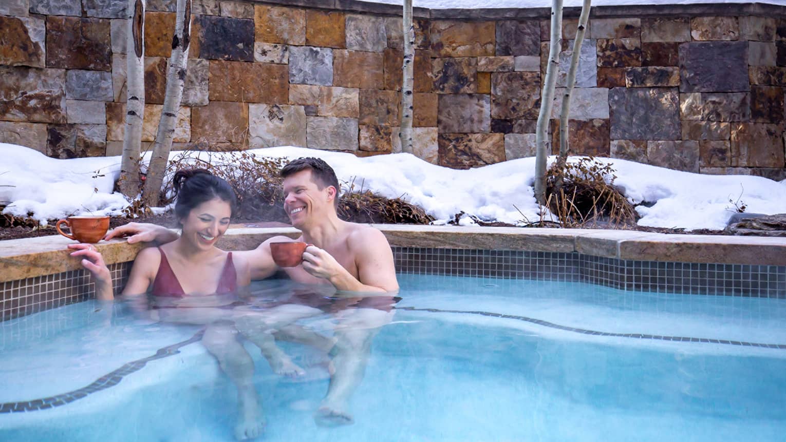 A woman and man drinking hot chocolate while sitting in a heated pool surrounded by snow.