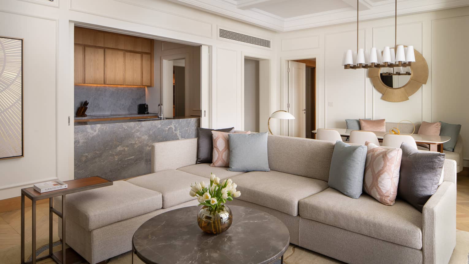 Contemporary living room with a grey sectional sofa, marble coffee table, open kitchen with wood cabinets and a dining area with pastel cushions.
