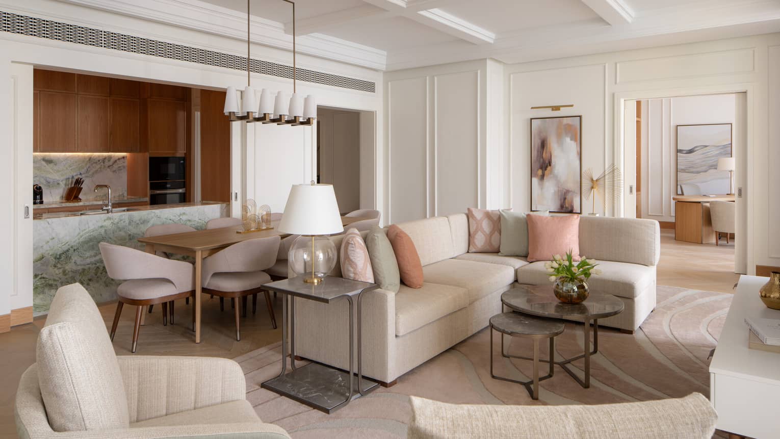 Elegant open-plan living space with neutral furnishings, pastel accents, a marble kitchen island and a dining area beside a modern chandelier.