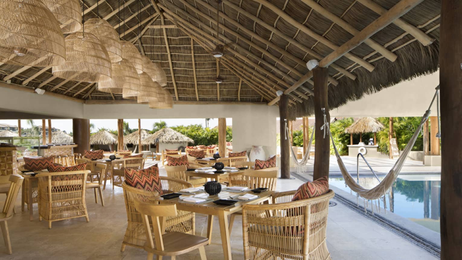 Whicker chairs and dining tables under thatched-roof pavilion, next to pool