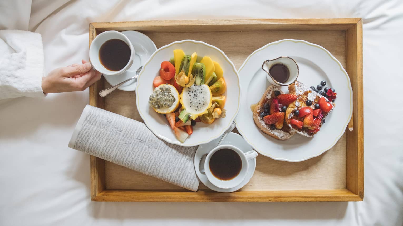 A wooden tray with various fruit, French toast and maple syrup served on white scalloped plates and two cups of coffee 