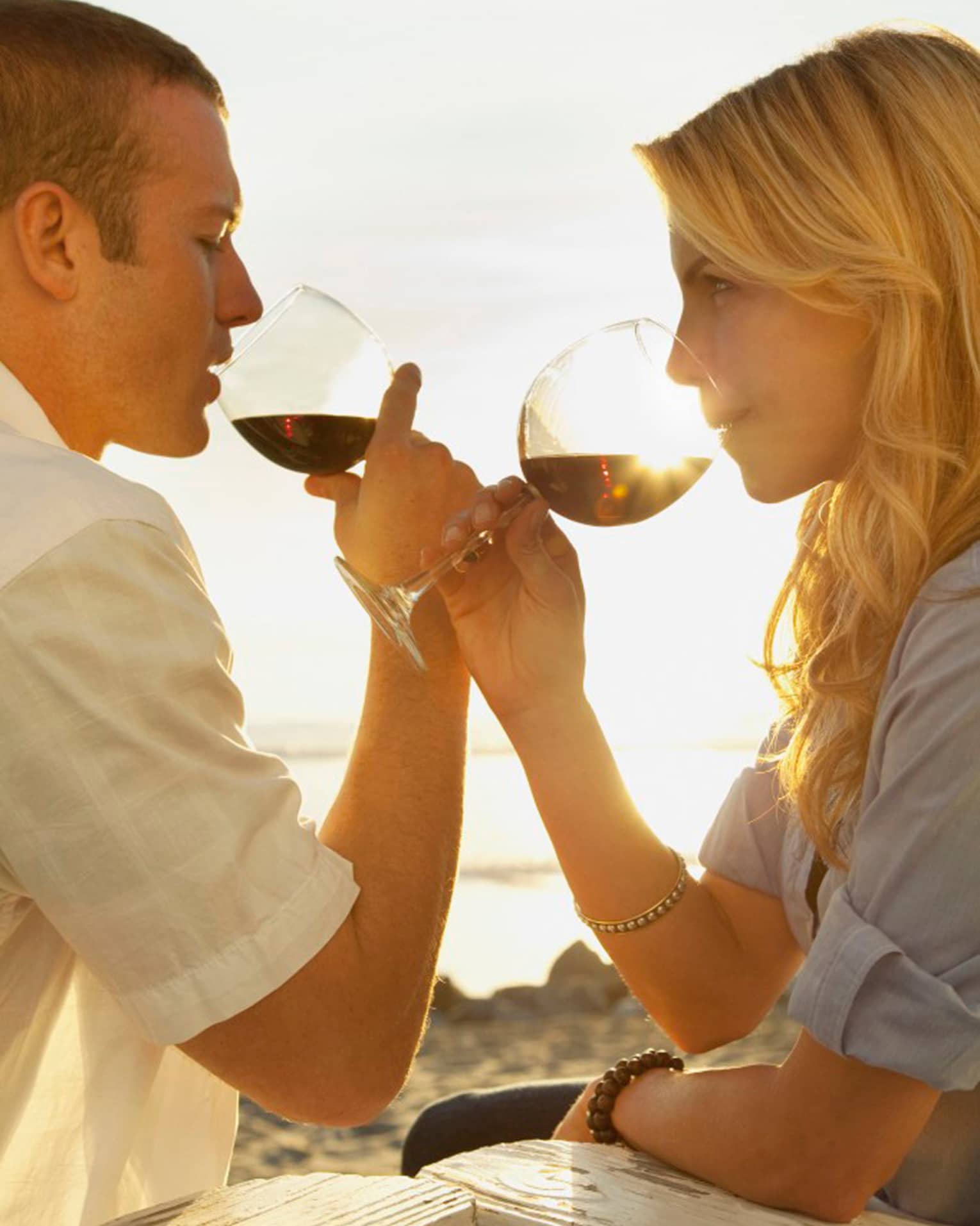 Couple sips red wine in large glasses, sun shining behind them
