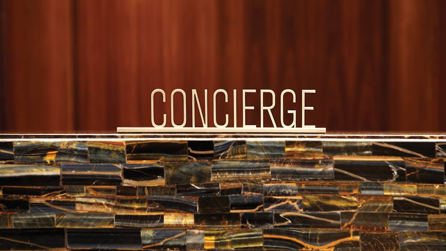 A metal concierge sign sites on top of stone reception desk in front of a wood paneled wall