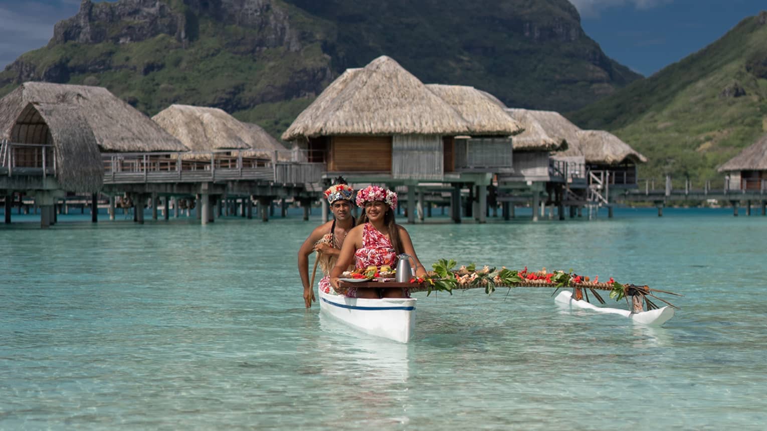 Two vibrantly dressed people in a canoe proffer a tray with colourful food attached to a pole draped with leaves and flowers.