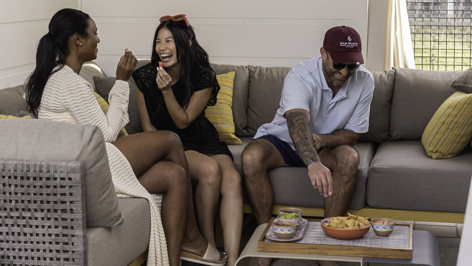 Three smiling guests eat tortilla chips with various dips while sitting on a sectional couch covered in colourful pillows.