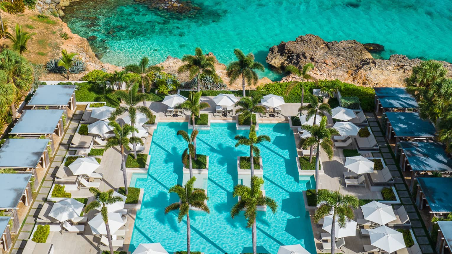 Aerial view of a pool surrounded by lounge chairs, palm trees and cabanas; beyond lies an expanse of turquoise ocean.
