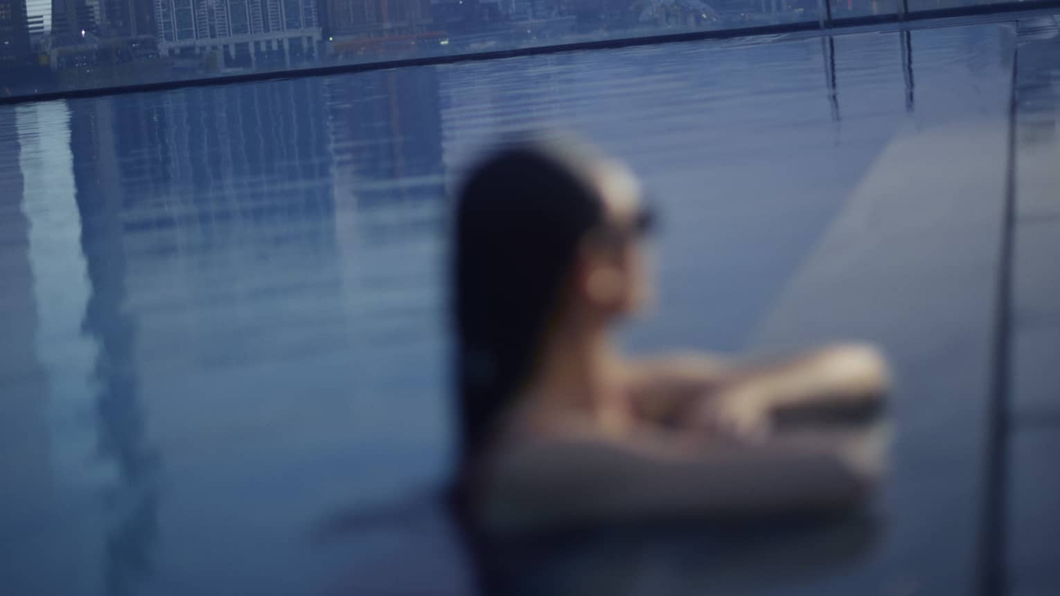 A woman in pool relaxes with elbows on pool deck, overlooking a city at dusk