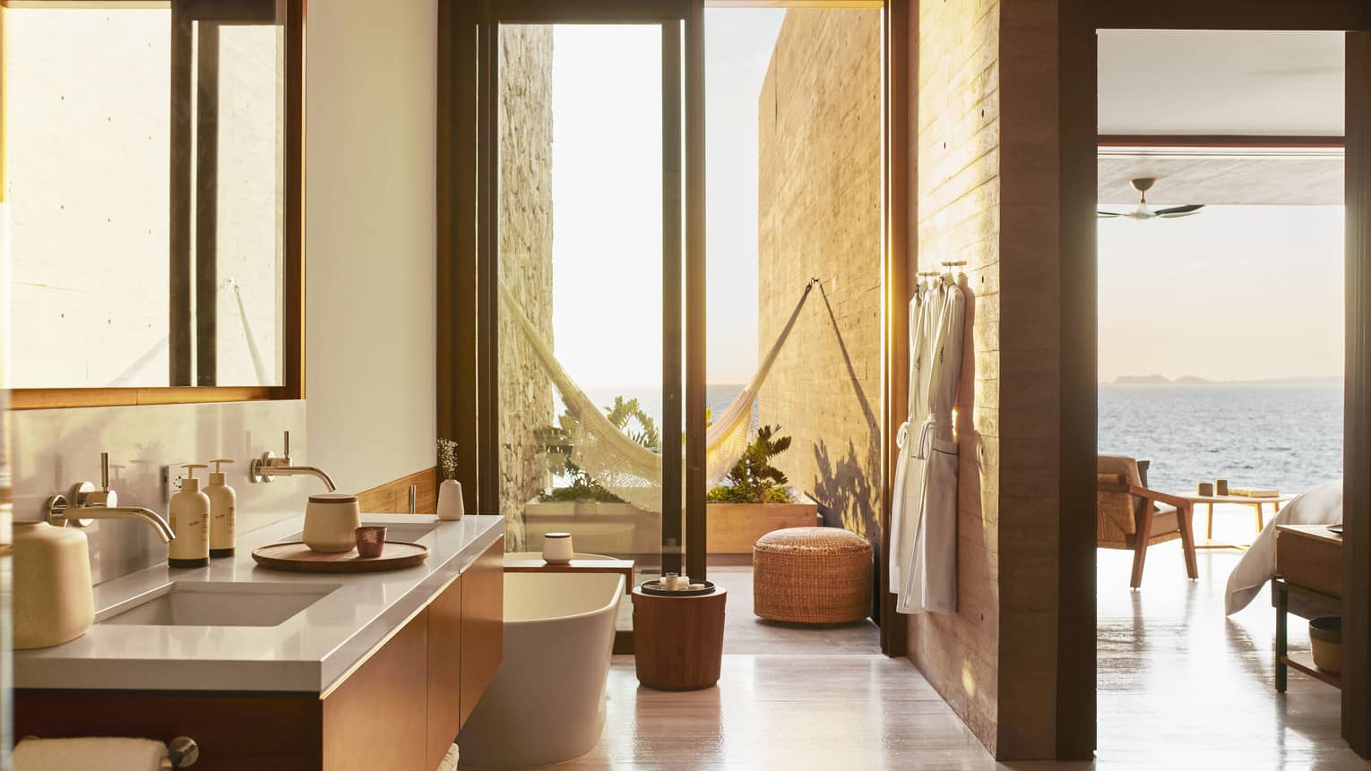 A luxurious bathroom with a double-sink vanity and free-standing tub, leading to a private patio with a hammock. The space opens to a scenic ocean view beyond.