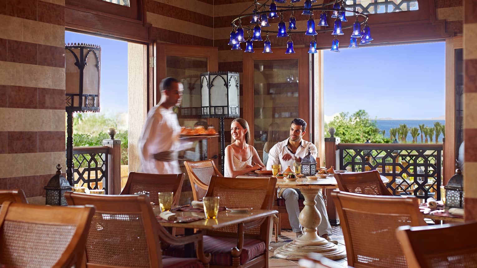 Couple sits at Arabesque corner table under blue chandelier, server walks by with tray