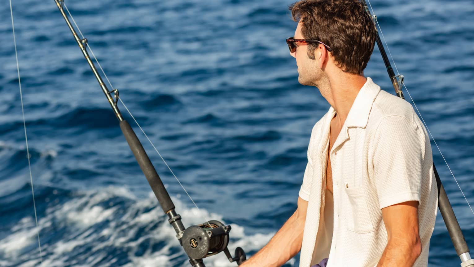 Close-up of a guest wearing sunglasses, sitting beside two offshore fishing rods and looking out towards dark blue waves.